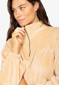 Pullover beige in corduroy con zip, colletto alto, texture a coste, polsini elastici e logo ricamato bianco sul petto.