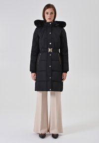 Zwarte gewatteerde jas met een capuchon van nepbont, een tailleband, drukknoopsluitingen en zakken met ritssluiting, gecombineerd met beige broek met brede pijpen.