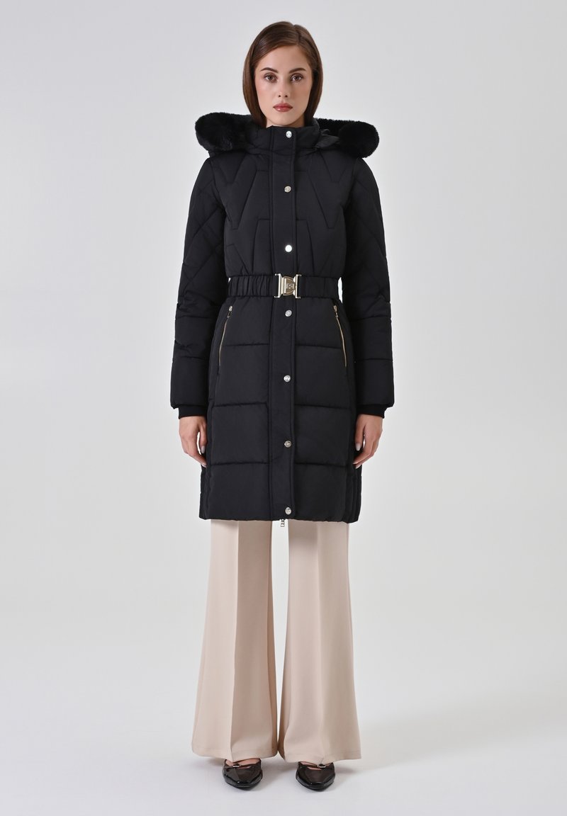 Zwarte gewatteerde jas met een capuchon van nepbont, een tailleband, drukknoopsluitingen en zakken met ritssluiting, gecombineerd met beige broek met brede pijpen.