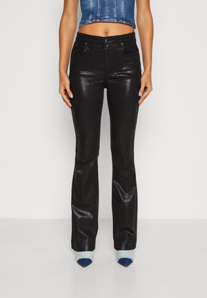 Kobieta w czarnych, błyszczących, rozszerzanych spodniach z wysokim stanem, krótkim niebieskim topie denimowym i butach na szpic z niebiesko-białym wzorem.