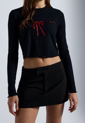 Kobieta ubrana w czarny crop top z długim rękawem z czerwoną wstążką na wysokości piersi oraz czarną mini spódniczkę z asymetrycznym pasem.