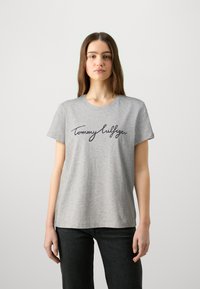 Camiseta de algodón gris con mangas cortas y cuello redondo, con un logotipo en cursiva negro "Tommy Hilfiger" en el frente.