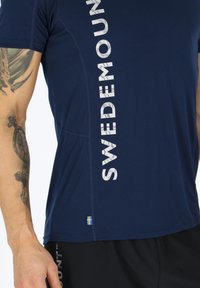 Marineblauw sportshirt met witte verticale lettering, korte mouwen en een klein accent van de Zweedse vlag op de zoom. Gladde stofstructuur.