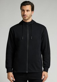 Sudadera negra con cremallera, de diseño estructurado, que incluye capucha con cordón, mangas largas y puños acanalados. Tejido suave y texturizado.