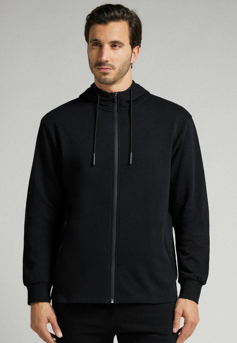 Sudadera negra con cremallera, de diseño estructurado, que incluye capucha con cordón, mangas largas y puños acanalados. Tejido suave y texturizado.