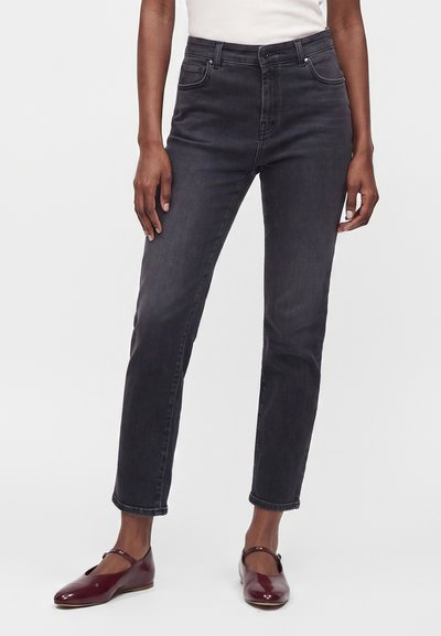 Jean gris foncé taille haute à coupe droite, porté avec un top blanc et des ballerines Mary Jane rouge foncé sur une personne debout.