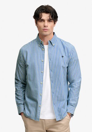 Mann trägt ein blau-gelb gestreiftes Hemd mit Knöpfen über einem weißen T-Shirt, beigen Hosen und hat die Hände in den Taschen, schaut nach vorne.