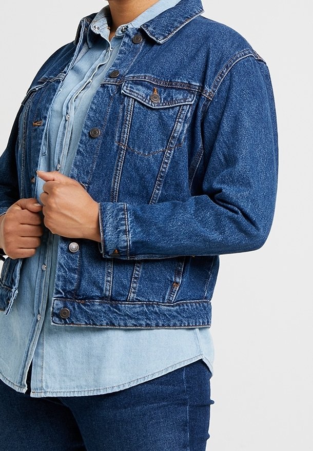 Chaqueta de mezclilla en azul oscuro con cierres de botón, bolsillos en el pecho y un dobladillo ligeramente recortado. Superpuesta sobre una camisa de mezclilla de color azul claro.