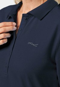 Marineblauwe polo met een kraag, twee knopen en een zilver geborduurd ontwerp op de linkerborst. De stof lijkt glad en gestructureerd.