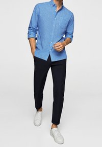 Camicia blu con bottoni e piccoli pattern bianchi, abbinata a pantaloni scuri e sneakers bianche. La camicia ha colletto e maniche lunghe.