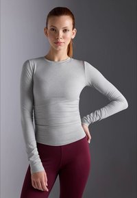 En ung kvinna med rött hår i en hästsvans, iklädd en tight grå långärmad tröja och vinröda leggings, som står mot en neutral bakgrund.