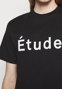 Personne portant un T-shirt noir à col rond avec un grand texte blanc "Étude" imprimé sur la poitrine sur un fond uni.