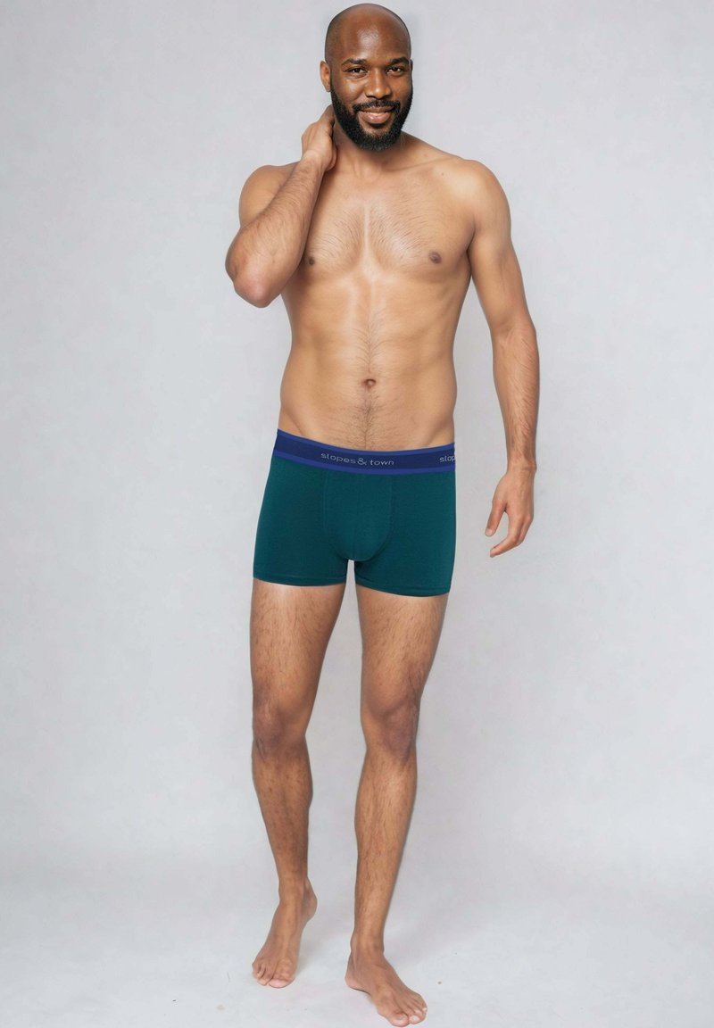 Męskie zielone bokserki z ciemnoniebieskim paskiem w talii, na którym widnieje nazwa marki. Materiał wydaje się być miękki o gładkiej fakturze.