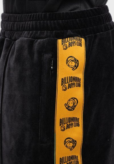 Μαύροι βελουτέ παντελόνες jogging με πορτοκαλί λωρίδα στο πλάι που φέρει το κείμενο και το λογότυπο "Billionaire Boys Club", περιλαμβάνοντας μια τσέπη με φερμουάρ στο πλάι.