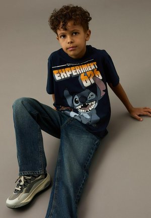 Jongen met krullend haar, gekleed in een marineblauw shirt met "Experiment 626" en een Stitch-illustratie, blauwe jeans en sneakers, zittend tegen een grijze achtergrond.