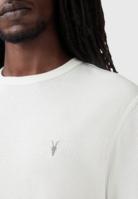 Vaaleanvihreä ribattu neulepaita pitkillä hihoilla, jossa on pyöreä pääntie ja pieni harmaa brodeerattu vuohikallon logo rinnassa.