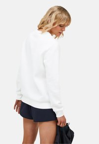 Felpa bianca a girocollo realizzata in tessuto morbido, con polsini e orlo a coste. Indossata con pantaloni corti scuri, presenta un design semplice e pulito.