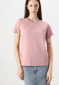 Vaaleanpunainen puuvillapaita lyhyillä hihoilla, jossa on kohotettu teksti "PEPE JEANS LONDON WEST ELEVEN" rinnassa. Pehmeä rakenne, rento muotoilu.