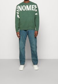 Pull en maille vert avec texte blanc "PUNOMEN", associé à un jean droit bleu clair et des baskets blanches. Look simple et décontracté.
