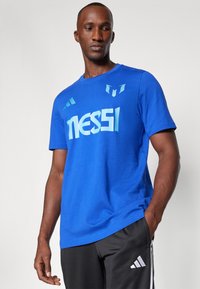 Blaues Baumwoll-T-Shirt mit "Messi"-Grafik in Hellblau, kurze Ärmel, runder Ausschnitt, kombiniert mit schwarzen Shorts mit weißen Adidas-Streifen.