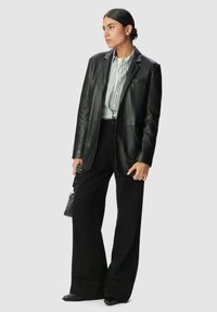 Blazer in pelle nera con una vestibilità sartoriale, abbinato a una camicia a righe con bottoni e pantaloni neri a gamba larga. Porta una piccola borsa testurizzata.