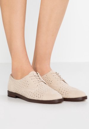Beige ruskind snørestøvler med perforeret design båret på bare fødder, kombineret med mørkebrune såler på et hvidt gulv.