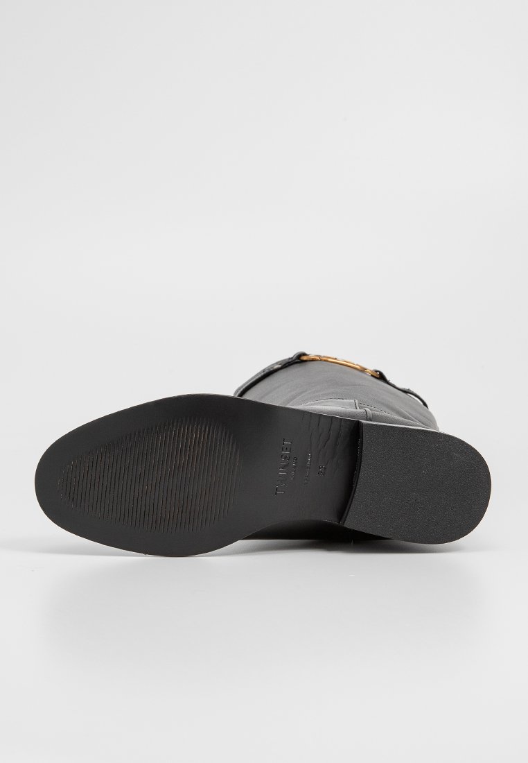 Stivale in pelle nera con punta arrotondata, suola in gomma caratterizzata da una texture orizzontale, e un accento decorativo dorato sulla cinghia vicino alla caviglia.