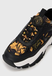 Black sneaker featuring gold and yellow floral patterns, leather and textile materials, prominent laces, and thick rubber sole.