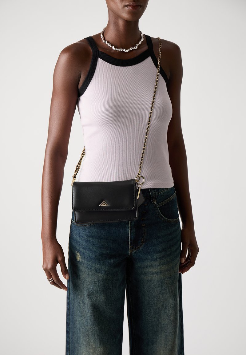 Black leather crossbody bag with a gold chain strap and triangular logo, held by a person wearing a light purple ribbed tank top and distressed jeans.