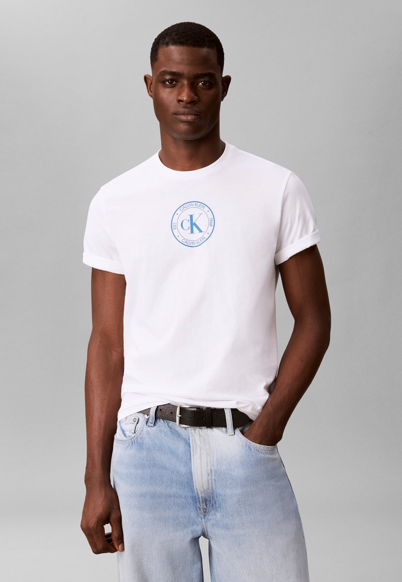 Άνδρας που φοράει λευκό T-shirt Calvin Klein με μπλε λογότυπο, ανοιχτό μπλε τζιν, μαύρη ζώνη, στέκεται μπροστά σε απλό γκρι φόντο.