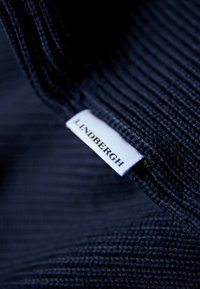 Tessuto a costine blu navy con un'etichetta bianca visibile che legge "LINDBERGH." La texture è morbida, con linee verticali ravvicinate.