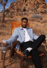 Lichtblauwe, lange mouwen shirt over een wit T-shirt, zwarte broek, zittend op een bruin leren stoel, met een glas ijskoud water in de hand.