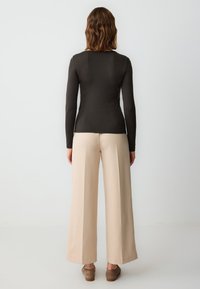 Maglione lungo a maniche nere di colore marrone scuro abbinato a pantaloni ampi beige, caratterizzati da una superficie liscia e una vestibilità rilassata, completati da scarpe marroni.