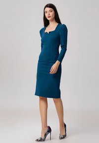 Robe ajustée bleu sarcelle avec décolleté carré, manches longues et accents de manches plissées. Associée à des chaussures à talons colorées.
