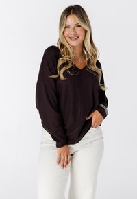 Femme jeune avec de longs cheveux blonds portant un pull ample foncé et un pantalon clair, souriant avec une main dans la poche devant un fond blanc.