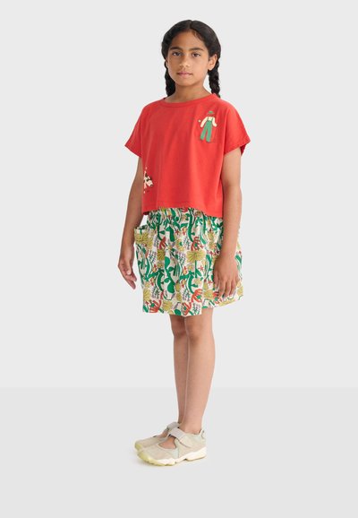 Fille aux cheveux tressés portant un t-shirt rouge avec de petites illustrations, une jupe fleurie et des chaussures beige à velcro sur un fond uni.