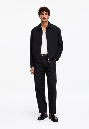 Homme debout portant une veste zippée noire, une chemise blanche, un pantalon noir avec une ceinture tressée et des mocassins en cuir noir sur fond blanc.