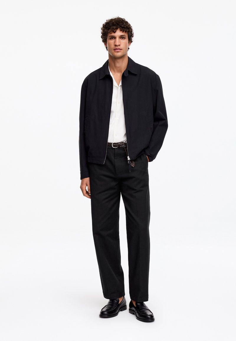 Homme debout portant une veste zippée noire, une chemise blanche, un pantalon noir avec une ceinture tressée et des mocassins en cuir noir sur fond blanc.