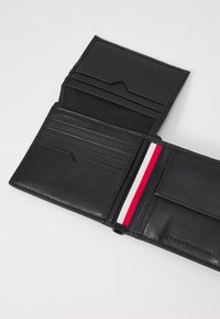 Portefeuille en cuir noir avec plusieurs emplacements pour cartes et un bandeau élastique central comportant des rayures rouge, blanche et bleu marine. Logo embossé présent.