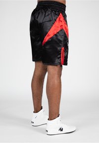 Zwarte satijnen sportshorts met een rood chevrondesign, elastische tailleband en zijsleuven. Gedragen met witte hoge sneakers.