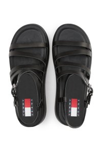 Sandales noires avec plusieurs brides plates, une semelle rembourrée et un patch logo présentant des couleurs rouge, blanc et bleu sur la semelle intérieure.