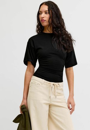 Žena s dugom kovrčavom tamnom kosom koja nosi pripijenu crnu majicu kratkih rukava i kremaste hlače s vezicama, držeći zelenu jaknu u jednoj ruci.