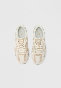 Pāris bēša un balta New Balance skriešanas apavu ar šņorēm, redzami no augšas uz gaiši pelēka fona.