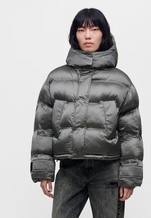Modèle portant une doudoune matelassée grise et brillante, avec une capuche oversize et un jean foncé, sur un fond clair uni.