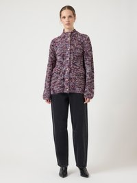 Grober Strickcardigan in einem mehrfarbigen Muster mit hohem Kragen, ausgestattet mit Knöpfen und zwei Fronttaschen. Getragen zu schwarzen Hosen mit weitem Bein.