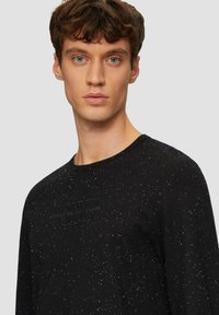 Schwarzes Langarmshirt mit einer gesprenkelten Textur und geprägtem Logodetail in der Mitte. Klassischer Rundhalsausschnitt und normale Passform.