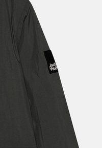 Giacca grigia scura con una texture liscia; la manica sinistra presenta una toppa in tessuto nera con il testo bianco "Jack Wolfskin."