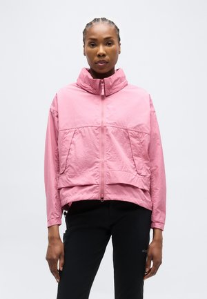 Žena s copánky, oblečená v růžové bundě s vysokým límcem na zip a černých kalhotách, stojící před jednobarevným bílým pozadím.