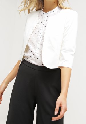 Femme portant un blazer court blanc par-dessus un chemisier blanc à pois et un pantalon noir taille haute, posant avec les bras détendus.
