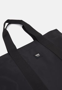 Black tote bag made of durable fabric with strong, flat handles. Features a small brand label on one side, no visible patterns or accents.